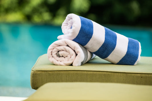 Two rolled-up towels, one blue and white striped and one pink, on a green cushion by a pool.